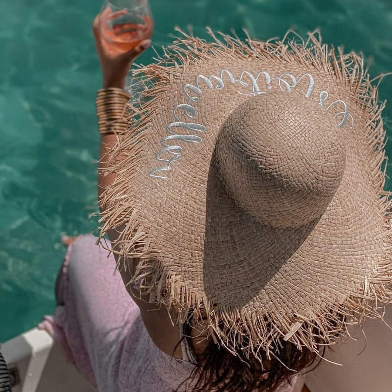 Person wearing a straw hat with 'Beach Life' text, holding a glass of pink wine by a pool.
